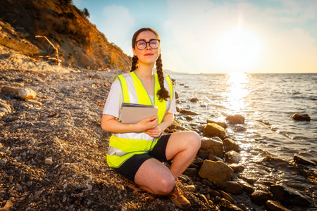 Earth day. Smiling woman wearing vest and holds electronic tablet squatting on beach near sea. Concept of ocean's pollution and protection of environment.の写真素材