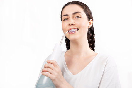 Portrait of young smiling woman with brackets on teeth cleans them using oral irrigator. White background. Concept of dental care during orthodontic treatment.の写真素材