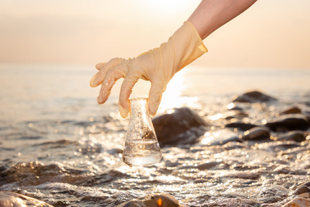 Close-up of hand in rubber glove holding glass flask with sample of pure ocean water. In background coast and sea. Concept of Earth Day and environmental protection.の写真素材