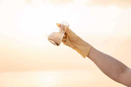 Close-up of hand wearing rubber glove holding flask with sample of dirty water. Sunset sky on background. Copy space. Concept of recycle water resources and ecology.の写真素材