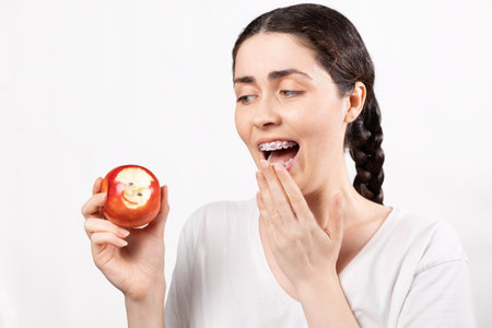 Portrait of shocked woman with brackets shows red apple with lost braces. White background. Concept...