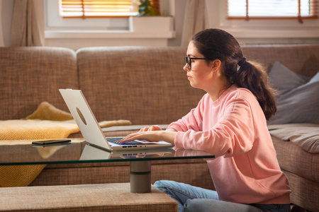 Side view of young Caucasian woman wearing glasses is sitting near sofa and using laptop. Concept of remote work and freelancing.の写真素材