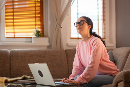Pensive young Caucasian woman wearing glasses is sitting on couch and using her laptop. Cozy interior. Concept of remote work and freelancing.の写真素材