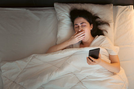 Top view of young Caucasian woman using smartphone and yawning, lying in bed at night. Insomnia, sleeplessness and social media addiction.の写真素材