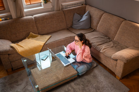 Top view of young Caucasian woman wearing glasses is sitting near sofa and using laptop. Concept of cozy remote work.の写真素材