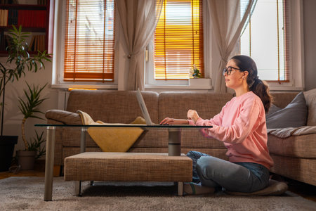 Side bottom view of young Caucasian woman wearing glasses is sitting near sofa and using laptop. Concept of cozy remote work.の写真素材