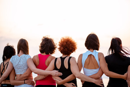Back view of group of fit women in sportswear stands in row and hugs each other. Light sunset sky in...