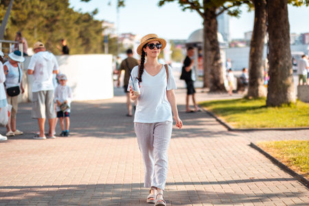 Young beautiful elegant Caucasian woman wearing straw hat and sunglasses walking at the city boulevard holding drink in her hand. Concept of summer season vacation.の写真素材