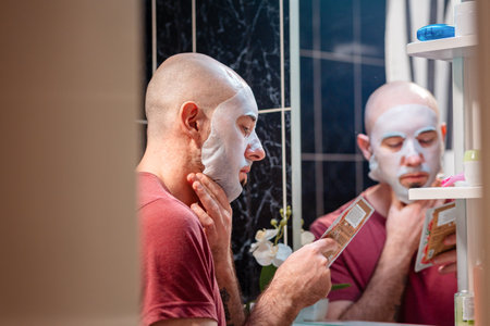 View from door at young adult Caucasian bald bearded man using cloth mask to moisturize the skin of his face. Beauty routine and skin care concept.の写真素材