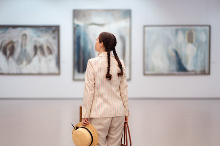 Back view of young Caucasian woman in elegant jacket and holds straw hat posing in the art gallery....