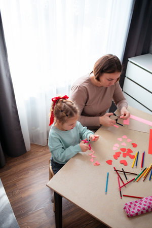 Top view of little cute girl and her mother cut out paper hearts sitting at a desk. Family is preparing for the celebration of Valentine's Day. Copy space.の写真素材