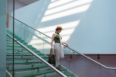 Wide view of a Caucasian pretty young elegant woman in a straw hat and carrying a bag descending transparent glass steps. Sunbeams at the wall. Copy space.の写真素材