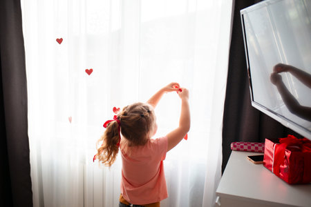 Rear view of little Caucasian girl decorate the curtains on the window with red paper hearts. A celebration for Valentine's Day and Mother's Day. Copy space.の写真素材