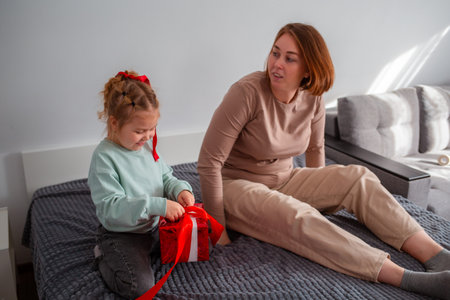 Mid shot of young Caucasian mother and her daughter pack a festive gift, sitting on the bed. The...