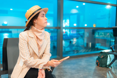 Mid shot of young Caucasian elegant woman is sitting in an airport lounge chair and holds smartphone. Windows in background. Concept of flight app.の写真素材