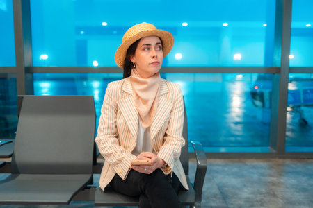 Mid shot of young elegant woman is sitting in an airport lounge chair and holds smartphone. Windows in background. Concept of flight and travel.の写真素材