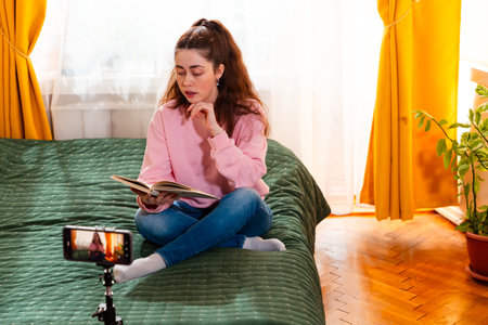 A stylish young woman read book and films a video while sitting on her bed, sharing engaging content for her social media followers. Mid shot.の写真素材