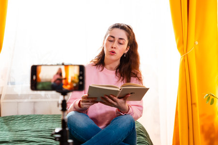 Caucasian woman sitting on a bed and making a video for her social media channel, using a tripod-mounted smartphone to communicate with her audience. Mid shot.の写真素材