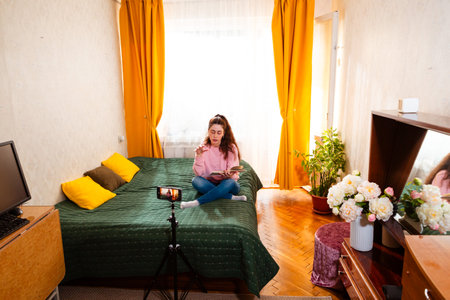 Young female entrepreneur recording a video on a smartphone tripod while sitting on her bed, engaging her audience in a vlog. Wide shot.の写真素材