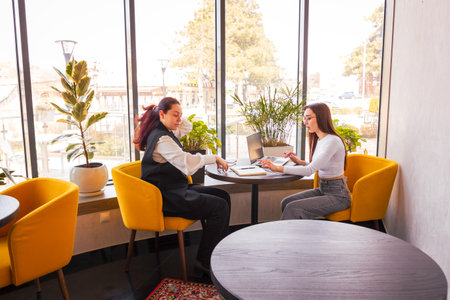 Business-savvy young women enjoying drinks and creative planning at a cafe. Collaboration over coffee. Concept of female-led entrepreneurship and mutual inspiration. Wide shot.の写真素材