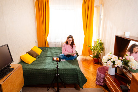 Young Caucasian woman sitting on a bed recording a video with a smartphone on a tripod, creating online content for social media and digital business. Wide shot.の写真素材