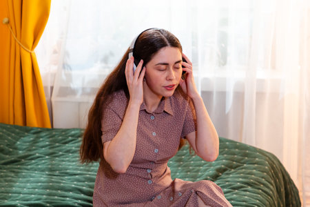 Young Caucasian woman sitting on a bed wearing headphones and listening to music at home. Mid shot.の写真素材
