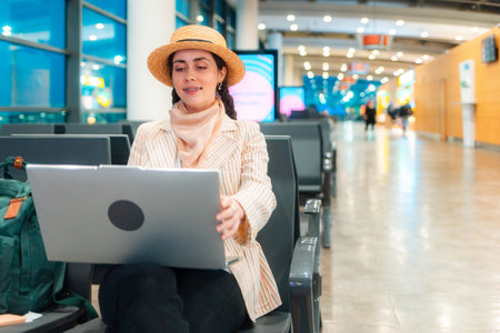 Mid shot of Caucasian elegant happy young woman is sitting on a chair in the waiting room of the airport and working on a laptop. Copy space. Concept of remote work and travel.の写真素材