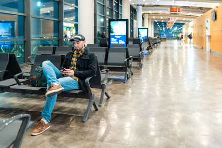 Mid shot of adult Caucasian man in cap and jacket using smartphone in airport waiting area sitting...