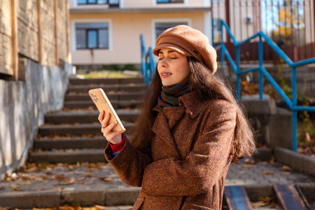Portrait of young pretty Caucasian happy woman in a coat and hat uses the phone while standing on...