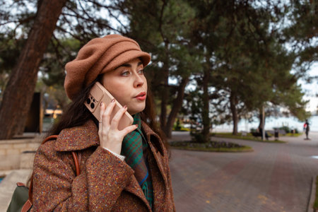 Portrait of a beautiful young woman in a hat and coat walking down the street and talking on the...