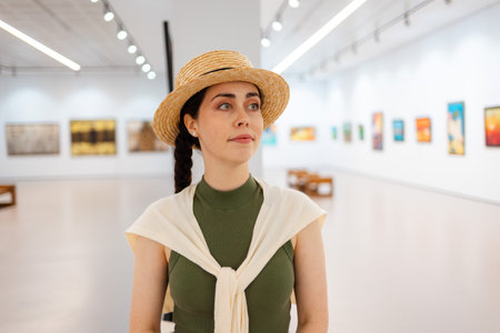 Portrait of a young elegantly dressed pretty woman student in a straw hat admiring the exhibition of...