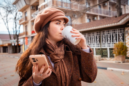 Portrait of stylish brunette woman enjoys a peaceful autumn walk along a city street while sipping...