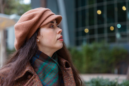 Portrait of a young Caucasian woman in a hat and scarf standing outdoors near a glass building....