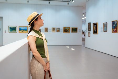 Mid shot of young Caucasian elegant woman in suit and straw hat contemplating pictures in the modern...