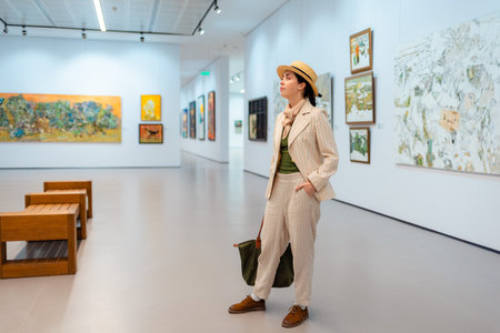 Museum day. Wide shot of young Caucasian pretty woman in elegant suit and straw hat posing in the art gallery. Concept of cultural education.の写真素材