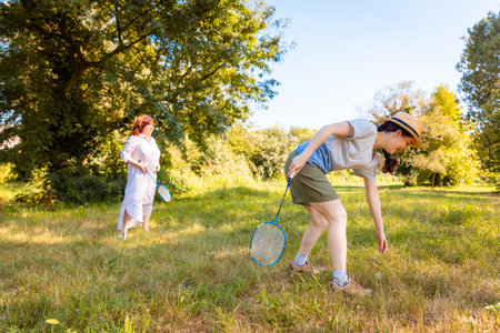 Caucasian mother and daughter dressed in casual clothes laughing while playing badminton in the city park. Family holiday, fitness and connection outdoorsの写真素材