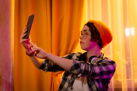 Portrait of casual teen girl wearing hat and headphones takes selfie on her bed in soft sunset light. Youthful lifestyle and personal digital time. Mid shot.の写真素材