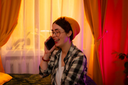 Portrait of girl talking on mobile while sitting on bed in evening light. Concept of emotional...