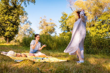 Summer picnic in the woods with two women, mom and daughter, talking and relaxing on a yellow blanket. Wide shot, low angle view.の写真素材