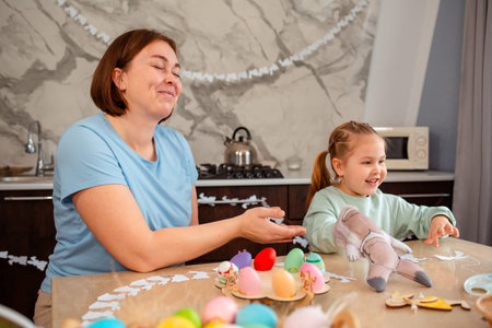 Smiling mother and happy little girl play together with rabbit glove, sitting at Easter table....