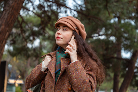 Portrait of a pretty young Caucasian woman in a hat and coat walking down the street and talking on...