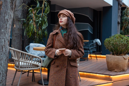 Young woman in a coat and beret walks through city streets while texting on her smartphone and...