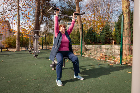 Older Caucasian woman using outdoor fitness machines in a green park. Physical activity, health, and...