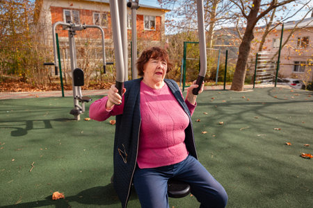 Senior Caucasian woman recovering after surgery exercises on outdoor fitness equipment in a park....