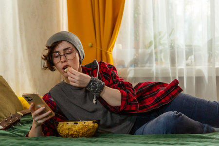 Caucasian teenage woman student resting indoors during free time. Girl eats popcorn and using smartphone. Home comfort theme. Mid shot.の写真素材