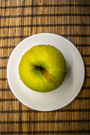 big ripe yellow green apple with brown line and water drops on a white small plate bamboo matの写真素材