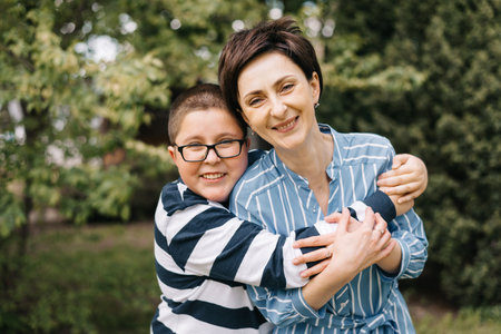 Mom with teenage son are walking in the park. They look at each other and smile. A woman hugs boy...