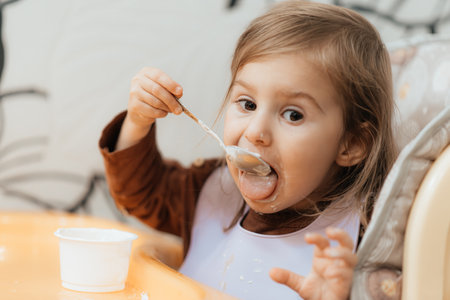 Small cute little toddler brunette caucasian girl with two tails tasting and enjoying by herself with a spoon the greek yogurt sitting in baby chair with messy face self-feeding conceptの写真素材