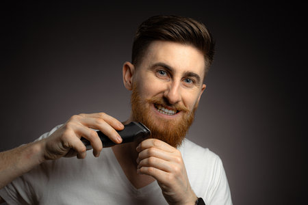 Handsome young bearded man trimming his beard with a trimmer