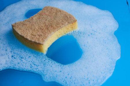 Yellow soapy sponge with foam on blue background. Cleaning concept. Copy spaceの写真素材